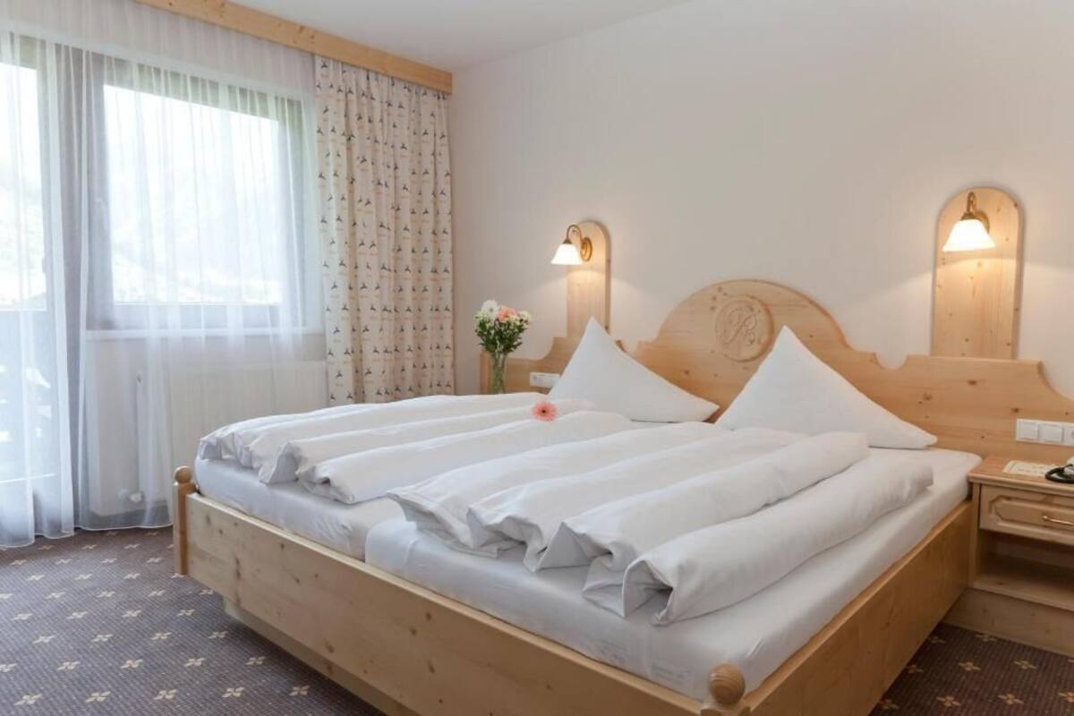 Un lit double avec du linge de lit blanc, une tête de lit en bois, des lampes de chevet, une table de chevet et une fenêtre avec des rideaux légers.