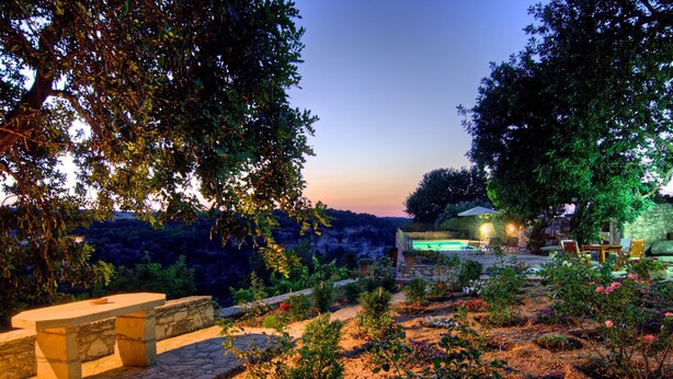 A stone bench and table set in a garden with a pool and patio furniture in the background. - Nature’s Peaceful Villa: Pool,BBQ, Fragrant Garden (Rethymno)