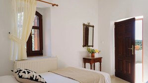 A bedroom with a bed, a window with curtains, a radiator, a wooden dresser with a mirror, and a door leading outside. - Nature’s Peaceful Villa: Pool,BBQ, Fragrant Garden (Rethymno)