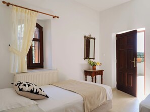 Ein Schlafzimmer mit Bett, einem Fenster mit Vorhängen, einem Heizkörper, einer Holzkommode mit Spiegel und einer Tür, die nach draußen führt.