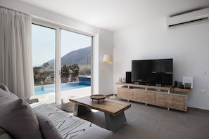 Ein modernes Wohnzimmer mit großem Fernseher, einem Holztisch und einer Couch. Eine Glastür führt zu einem Poolbereich mit Blick auf die Berge.
