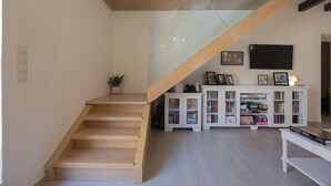 Eine Treppe führt zu einer Zwischenebene mit Glastrennwand, an der Wand ist ein Flachbildfernseher angebracht und ein weißes Unterhaltungselement mit Büchern und Gesellschaftsspielen ist vorhanden.