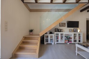 A staircase leading to a landing with a glass railing, a flat-screen TV mounted on the wall, and a white entertainment unit with books and board games. - Casa di Vangelis, Summer Village Stay! (Rethymno)