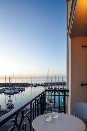 Ein Balkon mit Blick auf eine Marina bei Sonnenuntergang. Auf dem Tisch stehen zwei Gläser.