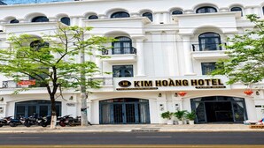 A white building with a sign reading 'Kim Hoang Hotel.' There are motorcycles parked in front, and a tree is visible. - KIM HOÀNG HOTEL TÂY NINH (Tan Ninh)