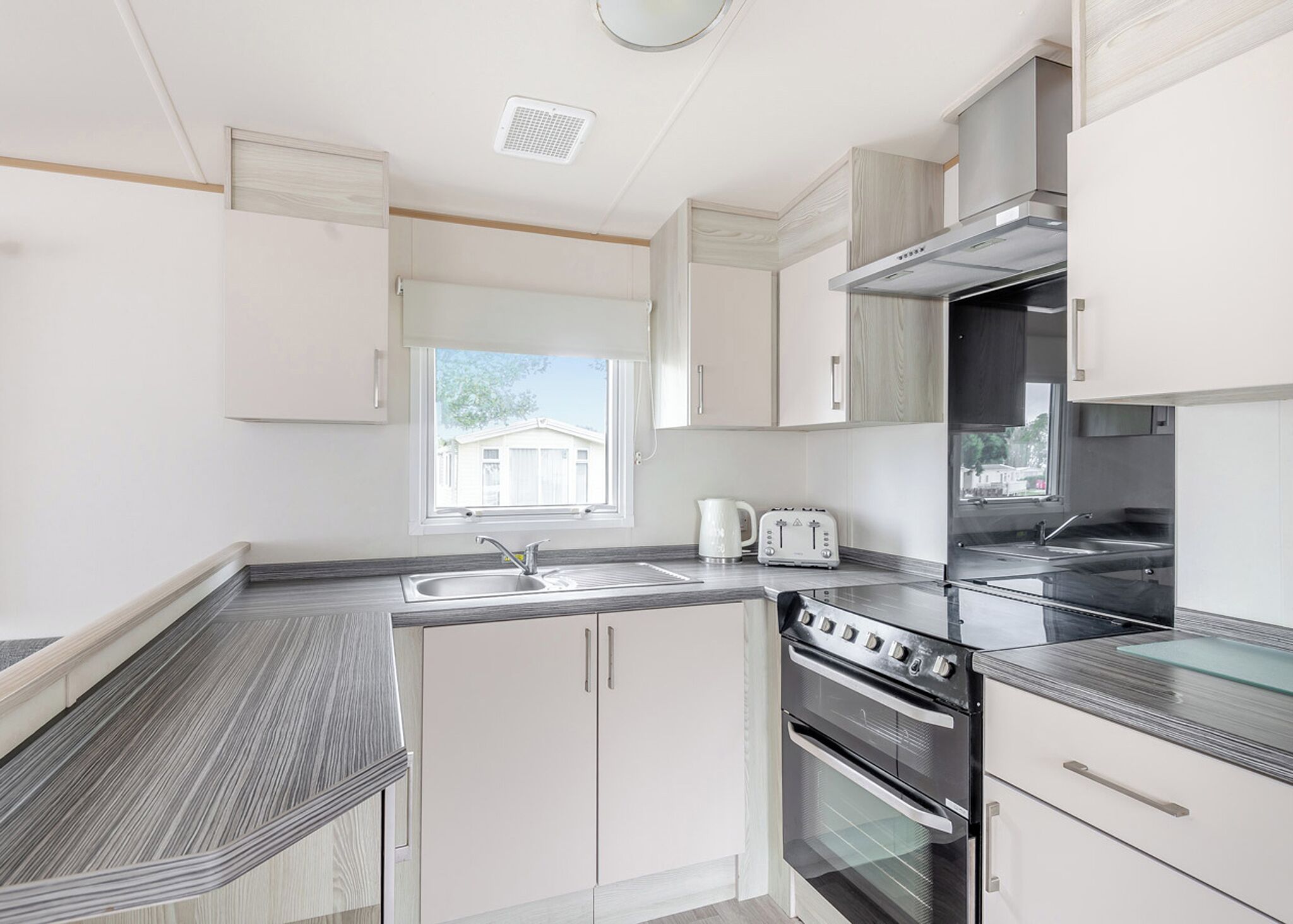 A modern kitchen with white cabinetry, a stainless steel range hood, a double oven, and a window with a view of a house.