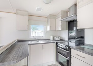 A modern kitchen with white cabinetry, a stainless steel range hood, a double oven, and a window with a view of a house. - Gold 4 Bedroom Caravan with parking and decking (Skegness)