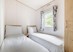 A bedroom with two single beds, a window with curtains, a wooden door, and a power outlet on the wall. - Gold 4 Bedroom Caravan with parking and decking (Skegness)