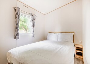 A bedroom with a bed, a window with curtains, a wooden headboard, and a nightstand. - Gold 4 Bedroom Caravan with parking and decking (Skegness)