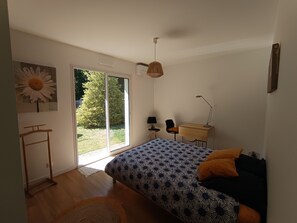 Ein Schlafzimmer mit einem Bett, einem Schreibtisch, einem Stuhl, einer Lampe, einem Fenster mit Blick ins Grüne und einer Tür.