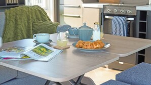 Eine Küchenzeile mit Herd, Backofen und Feuerlöscher. Ein Tisch mit einer Zeitschrift, einem Glas Saft, einem Teekessel und einem Teller mit Croissants.