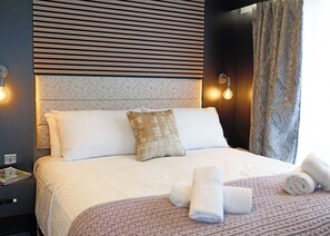 A neatly made bed with pillows, a bedside table with a magazine, and a wall with a patterned headboard and two wall sconces. - 2-bedroom S-Pod sleeps 4 (Landford, Nr Salisbury)