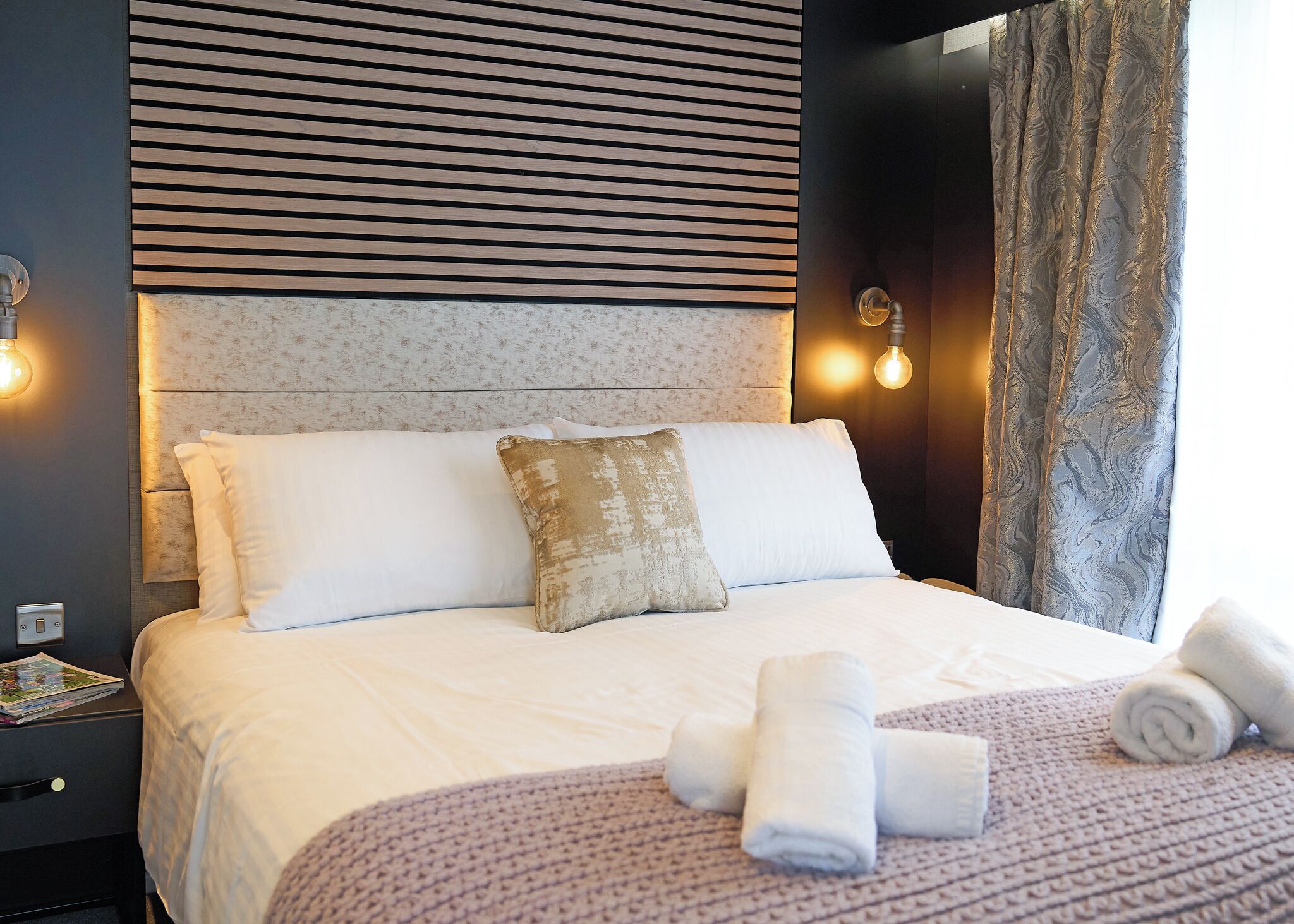 Una cama bien hecha con almohadas, una mesita de noche con una revista, y un cabecero decorado con un diseño y dos apliques de pared.