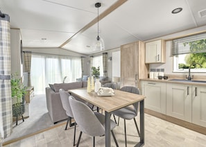 Une cuisine moderne avec un îlot central, un coin repas comprenant une table en bois et des chaises grises, et une vue sur la verdure par la fenêtre.