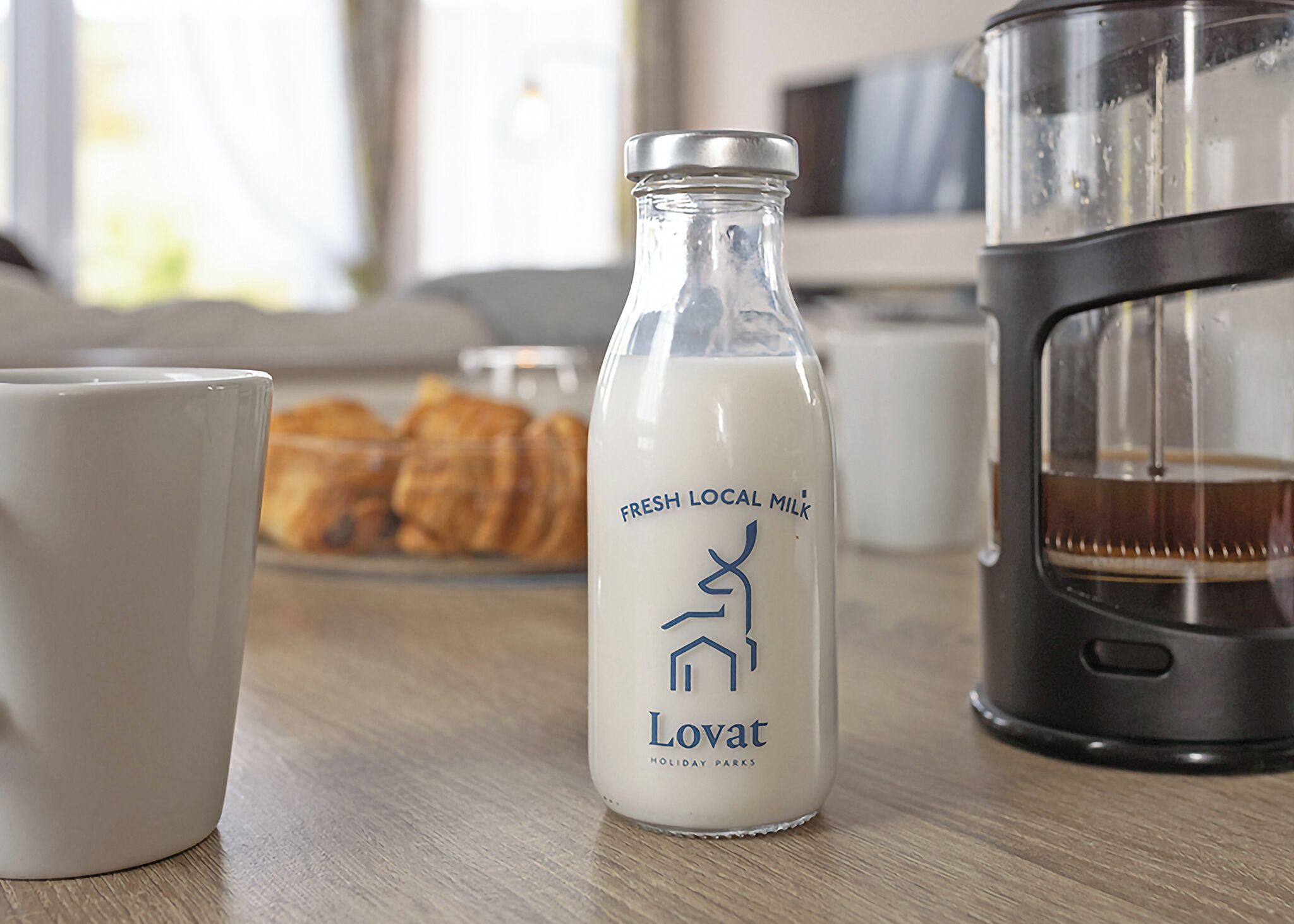 A bottle of fresh local milk, a white mug, and a coffee maker on a wooden table.