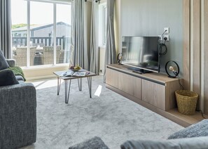 Un salon moderne doté d’un téléviseur à écran plat, d’une petite table avec des magazines, d’un panier en osier et offrant une vue sur un bâtiment à l’extérieur.