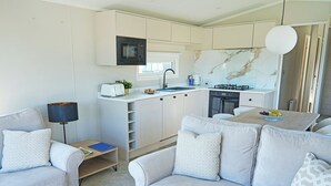 A modern kitchen with a built-in microwave, a gas stove, and a sink. There is a dining area with a table and chairs, and a living area with a sofa and a lamp. - Premium Hot Tub 4 bedroom Holiday Home sleeps 8 (Landford, Nr Salisbury)