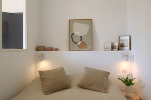 Une chambre à coucher avec un lit, des oreillers, un tableau abstrait accroché au mur, une petite étagère avec des objets décoratifs et un vase rempli de fleurs.
