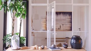 Ein Zimmer mit großem Fenster, einem Topfpflanzen, einer Glaskuppel, einem Gemälde und einem Holztisch mit diversen Gegenständen.