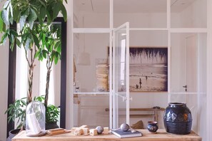 Ein Zimmer mit großem Fenster, einem Topfpflanzen, einer Glaskuppel, einem Gemälde und einem Holztisch mit diversen Gegenständen.