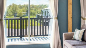 A room with a view of a green landscape, a balcony with a railing, and a couch with a cushion. - Exclusive 2 bedroom Sauna Lodge sleeps 4 (Landford, Nr Salisbury)