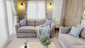 Ein Wohnzimmer mit einem beigen Sofa, einem Couchtisch, einem kleinen Tisch mit Lampe und einem Fenster mit Vorhängen.
