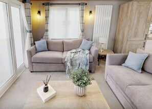 A living room with a beige sofa, a coffee table, a small table with a lamp, and a window with curtains. - Premium 2 bedroom Holiday Home sleeps 6 (Hayle)