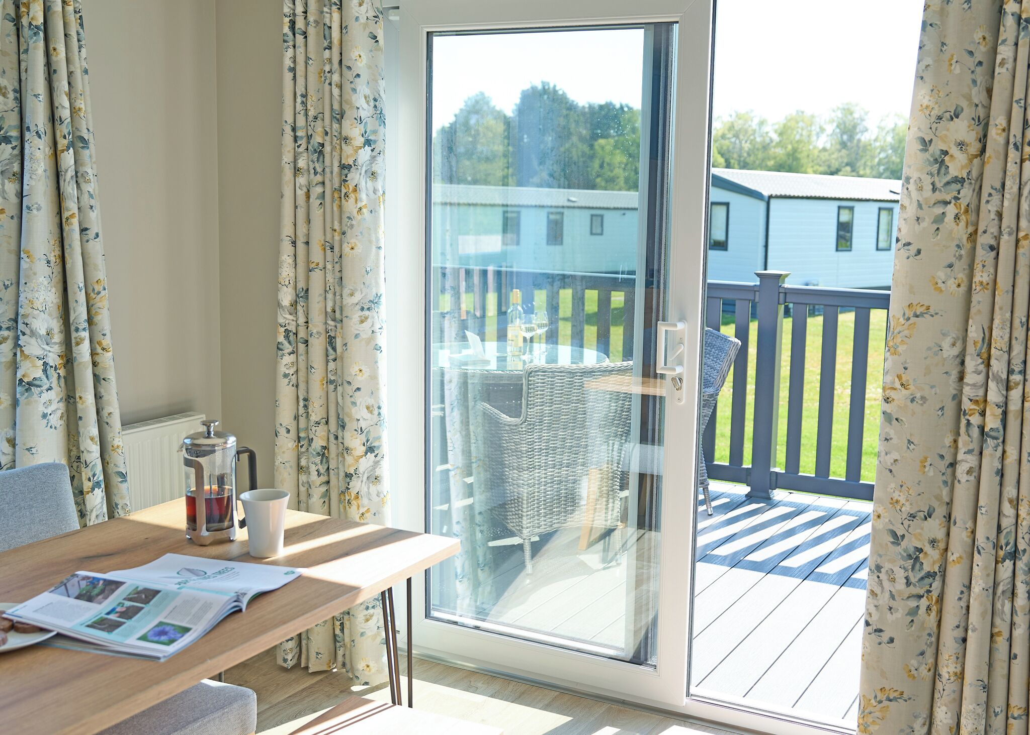 Une pièce avec une table en bois, une cafetière et un magazine. Une porte-fenêtre coulissante donne sur une terrasse extérieure aménagée avec des meubles en osier et offre une vue sur une maison.
