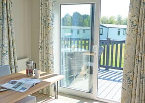 A room with a wooden table, a coffee pot, and a magazine. There is a sliding glass door leading to an outdoor deck with wicker furniture and a view of a house. - Exclusive 2 bedroom Lodge sleeps 4 (Pet) (Landford, Nr Salisbury)