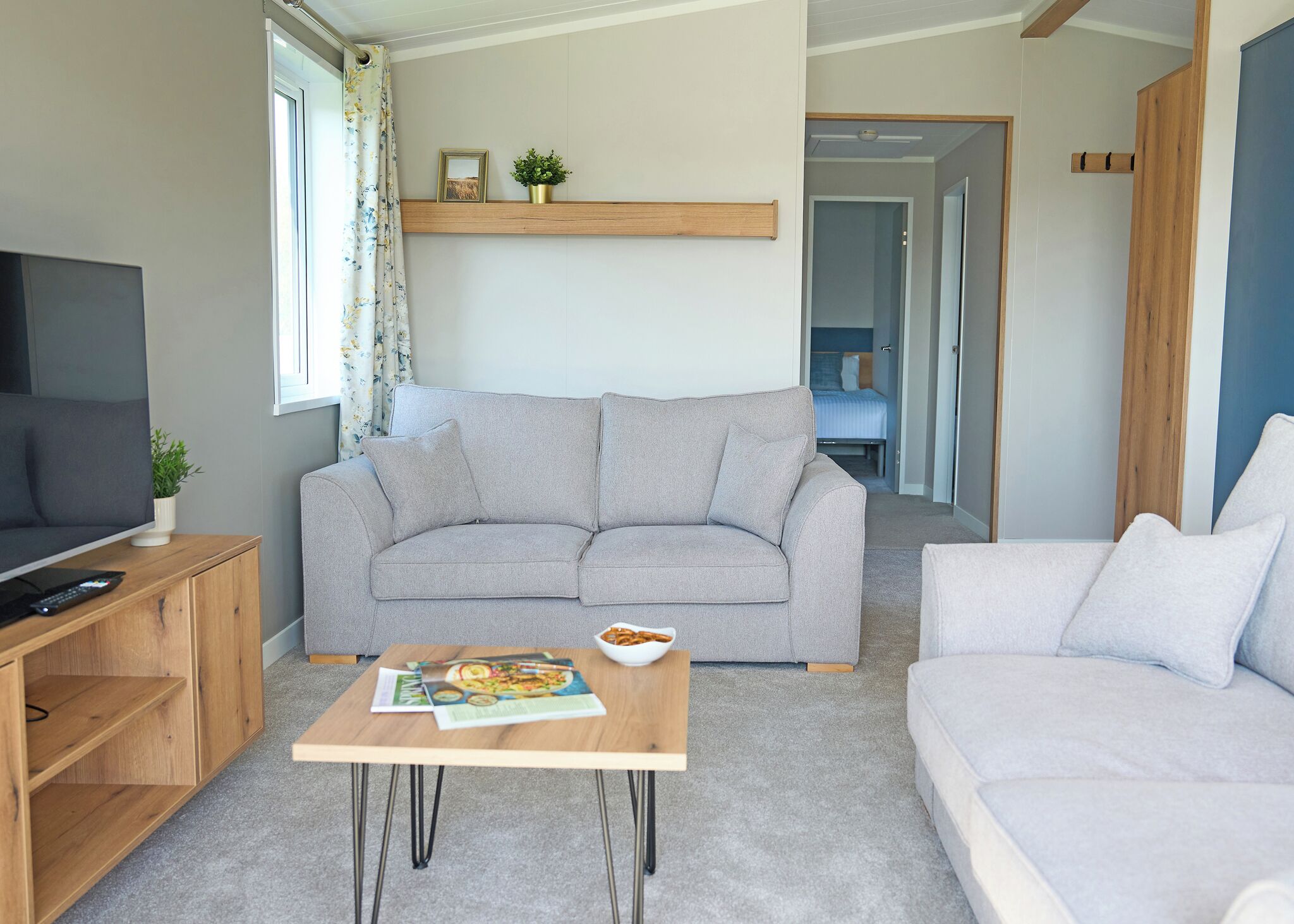 A modern living room with a grey sofa, a wooden coffee table with magazines, a TV on a wooden stand, and a small plant.
