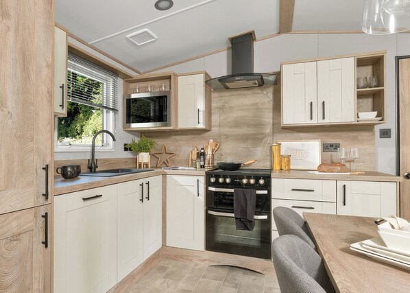 A modern kitchen with a built-in stove, oven, and microwave, wooden cabinets, and a window with blinds. - Premium Hot Tub 2 bedroom Holiday Home sleeps 6 (Hayle)