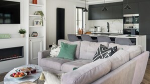 A modern living room with a grey sofa, a coffee table with fruits, a fireplace, and a kitchen area with dark cabinets and a microwave. - Autograph Kos Hot Tub 2-bedroom Luxury Lodge slp 4 (Landford, Nr Salisbury)