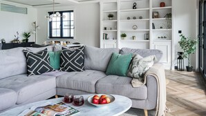 A modern living room with a grey sectional sofa, a round coffee table with magazines and fruit, and a white shelving unit with decorative items. - Autograph Kos Hot Tub 2-bedroom Luxury Lodge slp 4 (Landford, Nr Salisbury)
