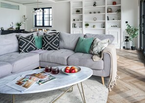 A modern living room with a grey sectional sofa, a round coffee table with magazines and fruit, and a white shelving unit with decorative items.