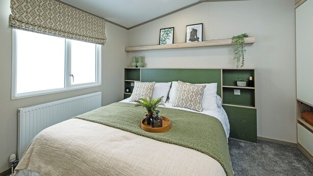 Een slaapkamer met een bed, een raam met een decoratieve gordijnboog, een radiator, een plank met decoratieve objecten, en een plant.