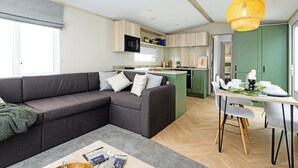 Een moderne woonkamer met een hoekbank, een eettafel met stoelen, een keukenhoek en een groot raam.