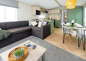 A modern living room with a sectional sofa, a dining table with chairs, a kitchen area, and a large window. - A stylish and contemporary retreat designed with comfort and quality in mind. (Peterborough)