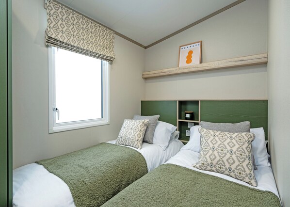 Quarto com duas camas, uma janela com valência estampada, cabeceira verde, uma prateleira com uma moldura e um cobertor verde em cada cama.