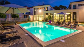 Ein Schwimmbecken mit Liegestühlen und Sonnenschirmen, umgeben von Gebäuden mit großen Fenstern und einer gut beleuchteten Fassade.