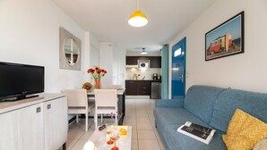Ein modernes Wohnzimmer mit Flachbildfernseher, einer Couch, einem Couchtisch mit Dekoration, einem Essbereich mit weißem Tisch und Stühlen sowie einer Küche mit dunklen Schränken und einer Mikrowelle.