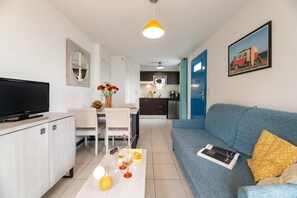 A modern living room with a flat-screen TV, a sofa, a coffee table with decorative items, a dining area with a white table and chairs, and a kitchen with dark cabinets and a microwave. - Seaside apartment in Saint-Palais-sur-Mer (Saint-Palais-sur-Mer)