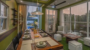 A dining area with a long table set for four, a ceiling fan, and large windows overlooking a garden. - ELIVAAS Moonlit retreat 3 BHK with Pool (Alibag)