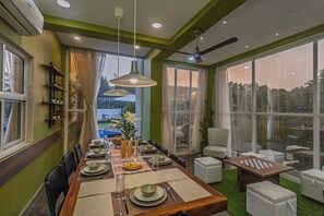 A dining area with a long table set for four, a ceiling fan, and large windows overlooking a garden. - ELIVAAS Moonlit retreat 3 BHK with Pool (Alibag)