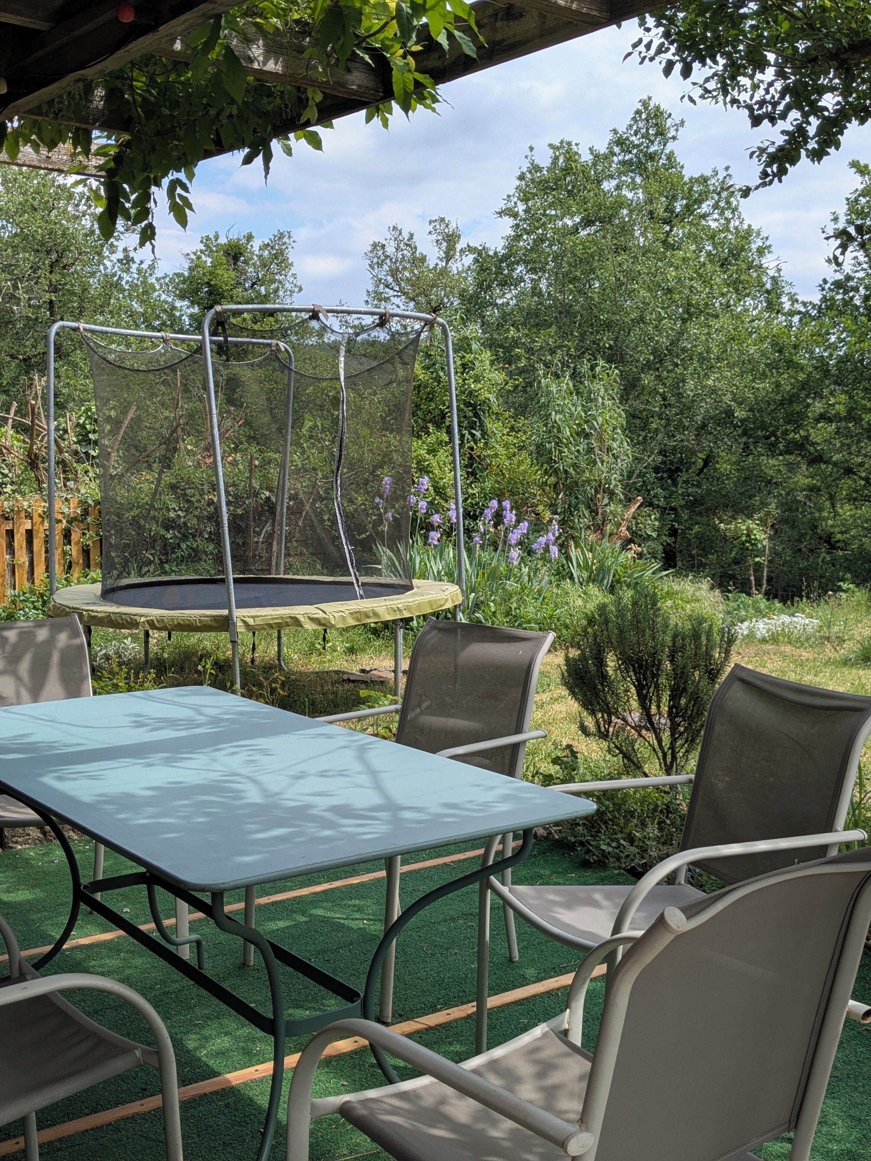 An outdoor patio with a table and chairs, a trampoline, and a garden with trees and plants.
