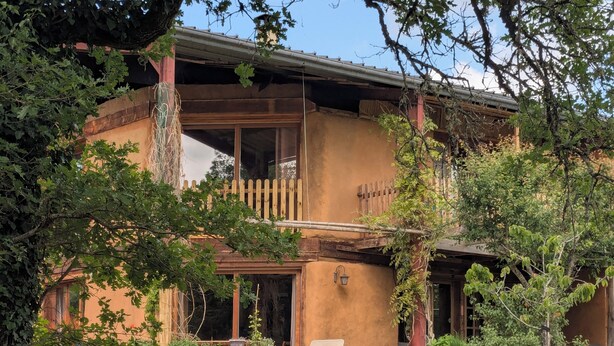 Un edificio rústico de dos pisos con balcón de madera, un porche con una silla y un jardín.