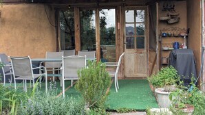 Un patio exterior con una pérgola de madera, una mesa con sillas, una puerta con paneles de vidrio y un estante con utensilios de cocina.