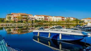 Uma marina com barcos atracados, um corpo d'água calma refletindo edifícios e um céu azul límpido.