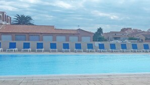 Piscina com espreguiçadeiras, um edifício com telhado de azulejos e palmeiras ao fundo.