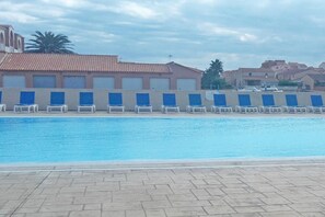 Ein Schwimmbecken mit Liegestühlen, ein Gebäude mit Ziegeldach und Palmen im Hintergrund.