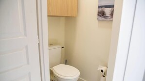 Et lille, hvidt badeværelse med toilet, et træskab over og et billede på væggen.